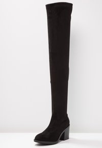 Stivale nero sopra il ginocchio in morbida ecopelle scamosciata, con tacco spesso e una texture liscia. Design elegante e alto con punta arrotondata.