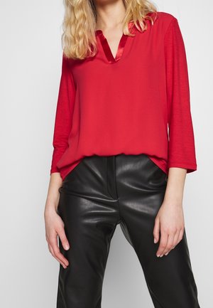 Femme portant une blouse rouge à manches longues avec un col en V en satin et un pantalon en cuir noir, debout les mains détendues le long du corps.