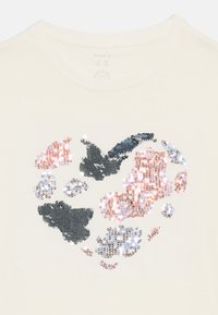 Cremefarbenes T-Shirt mit einem Herz aus mehrfarbigen Pailletten in Rosa-, Silber- und Marineblau-Tönen, gefertigt aus einem weichen, leichten Stoff.