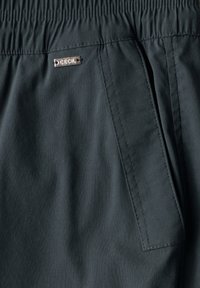Pantalons vert foncé avec une texture lisse, dotés d'une taille élastique et d'une poche latérale. Une petite étiquette de marque en métal est visible.