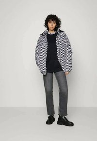 Casaco de chuva com padrão de ondas em preto e branco, com capuz, usado sobre uma blusa escura, combinado com jeans cinzentos e botas pretas com atacadores. Fundo simples.
