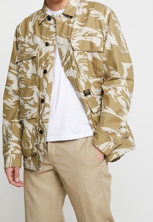 Personne portant une veste camouflage beige et blanche sur une chemise blanche et un pantalon beige avec une main dans la poche.