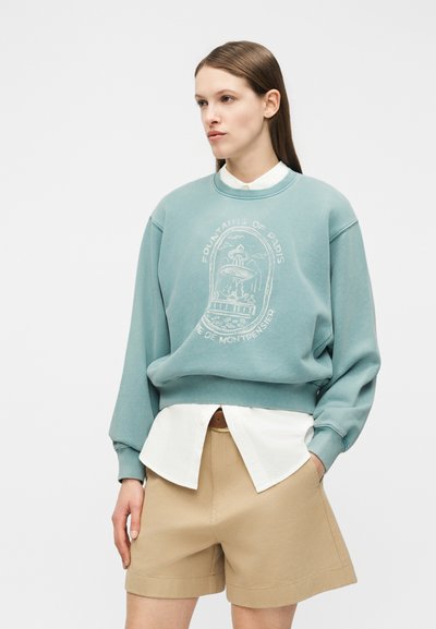 Γυναίκα που φοράει μπλούζα σε τιρκουάζ απόχρωση με το σχέδιο «Fontaines de Paris», λευκό πουκάμισο χωρίς να είναι μπερδεμένο από κάτω και μπεζ σορτς πάνω σε απλό φόντο.