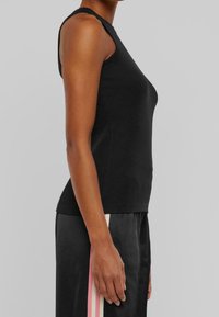 Černý top bez rukávů z hladké látky, s přiléhavým střihem a kulatým výstřihem. Nošený s černými kalhotami s růžovými pruhy.