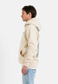 Sudadera color crema con un bolsillo frontal, diseño verde en el lado izquierdo y puños acanalados. Tela suave con un corte relajado.
