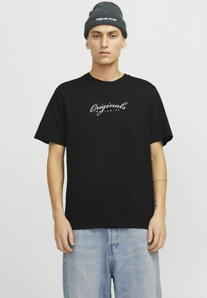 Camiseta de algodón negra con mangas cortas, que presenta un logo en forma de letra blanca en el pecho; combinada con unos vaqueros de mezclilla azul claro y un gorro gris.
