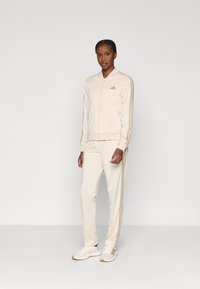 Μπεζ φόρμα από απαλό ύφασμα, που περιλαμβάνει μπουφάν με φερμουάρ και στενά παντελόνια με χρυσές ρίγες στα πλάγια. Φοριέται με λευκά αθλητικά παπούτσια.