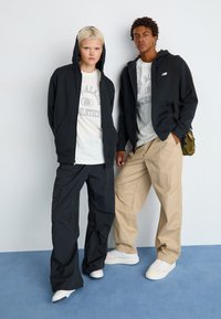 Des sweats à capuche zippés noirs sur des t-shirts graphiques clairs, associés à des pantalons cargo noirs et des pantalons larges beiges. Des baskets complètent les tenues.