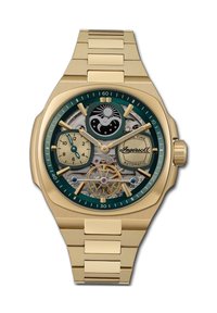 Goudkleurig automatisch horloge met een groene bezel, een skeleton wijzerplaat die de tandwielen toont, twee subwijzers en een gestructureerde gouden armband.