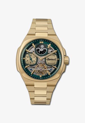 Goudkleurig automatisch horloge met een groene bezel, een skeleton wijzerplaat die de tandwielen toont, twee subwijzers en een gestructureerde gouden armband.