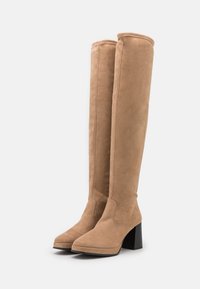 Bocanci din suede de culoare bej, până la genunchi, cu vârf pătrat și toc bloc. Textură netedă și design minimalist, cu ștecheri vizibile de-a lungul marginilor.