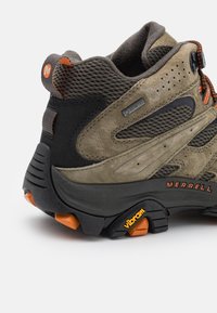 Brązowy zamszowy but trekkingowy z czarną siateczką i szarymi akcentami. Posiada pomarańczowe sznurówki i podeszwę Vibram. Zawiera etykietę Gore-Tex.