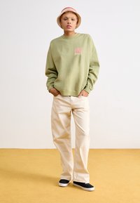 Olive green sweatshirt with pink graphic, paired with light beige wide-leg pants and black sneakers with white soles.