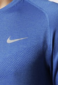 Niebieska koszulka sportowa wykonana z teksturowanej tkaniny, z charakterystycznym diamentowym wzorem po jednej stronie oraz srebrnym logo Nike na piersi.
