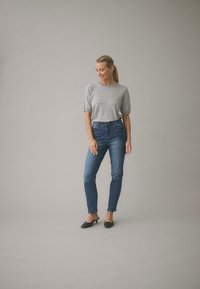 Grå randig kortärmad topp kombinerad med mörkblå slim-fit jeans. Svarta skor med en liten klack. Enfärgad grå bakgrund.