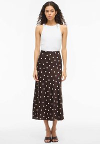 Brązowa spódnica midi w groszki o gładkiej fakturze. Połączona z dopasowanym, białym, bezrękawnym topem. Modelka stoi w płaskich sandałach.
