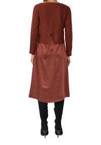 Frau in einem rostfarbenen satin Midi-Kleid, braunem, kurz geschnittenem Pullover mit einem Rückenschlitz und schwarzen, hochhackigen Kniefallstiefeln, von hinten betrachtet.