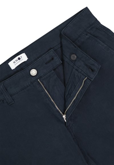 Pantalon bleu marine foncé avec fermeture éclair partiellement ouverte, fermeture par bouton et étiquette blanche indiquant "NN07 No Nationality" à l'intérieur de la ceinture.