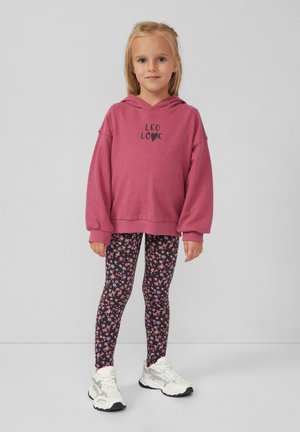 Ein junges Mädchen steht in einem rosa Kapuzenpullover mit dem Text "LEO LOVE", schwarzen Blumenleggings und weißen Turnschuhen vor einem schlichten grauen Hintergrund.