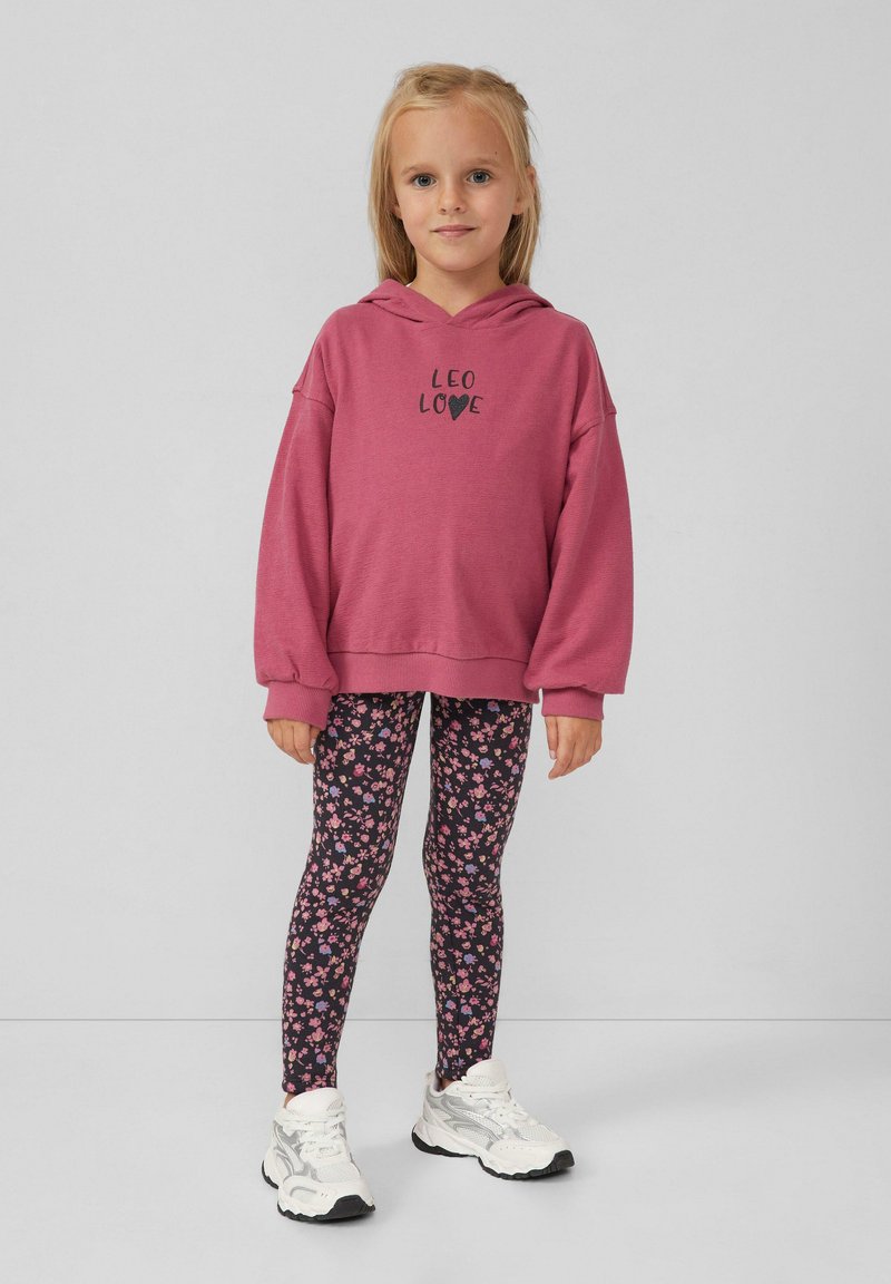 Ein junges Mädchen steht in einem rosa Kapuzenpullover mit dem Text "LEO LOVE", schwarzen Blumenleggings und weißen Turnschuhen vor einem schlichten grauen Hintergrund.