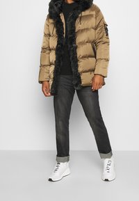 Chaqueta de abrigo marrón con capucha y cuello de piel sintética, combinada con jeans oscuros y zapatillas deportivas blancas. Cuenta con un bolsillo lateral con cremallera.