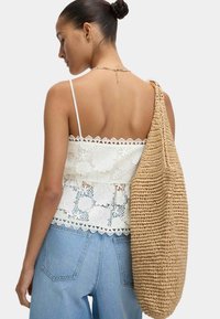 Weiße Spitze-Camisole mit floralen Mustern und gewelltem Saum, kombiniert mit einer beigefarbenen, gewebten Tasche. Blaue Jeans mit sichtbaren hinteren Taschen.