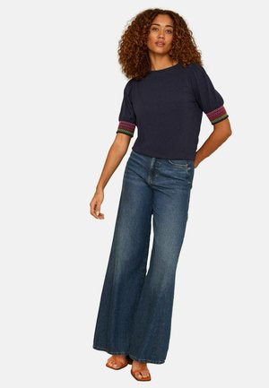 Femeie cu păr creț purtând un top bleumarin cu manșete colorate, blugi evazați albaștri și sandale portocalii, stând cu o mână pe șold.