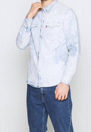 Mężczyzna w jasnoniebieskiej koszuli z denimu w stylu acid-wash z zatrzaskami i niebieskich dżinsach, dotykający kołnierzyka lewą ręką.