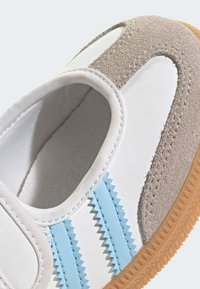 Biele a sivé tenisky s modrými cik-cak pruhmi, zamša akcentami a textúrovanou gumovou podrážkou. Zaoblený tvar, polstrovaný golier pre pohodlie.