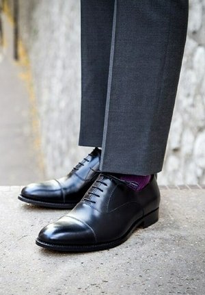 Zwarte leren dresschoenen met een cap toe-ontwerp, voorzien van een gladde afwerking en elegante veters, gecombineerd met donkergrijze pantalons en paarse sokken.