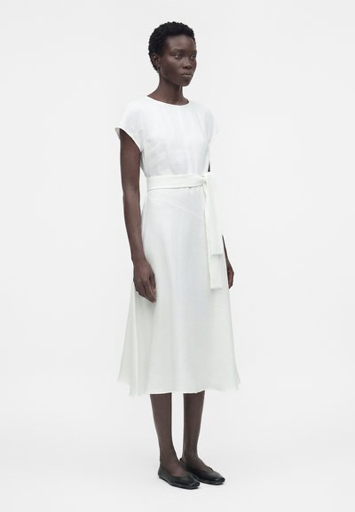 Γυναίκα που στέκεται φορώντας λευκό midi φόρεμα με κοντά μανίκια και δεμένη ζώνη στη μέση, σε συνδυασμό με μαύρα επίπεδα παπούτσια, σε απλό φόντο.