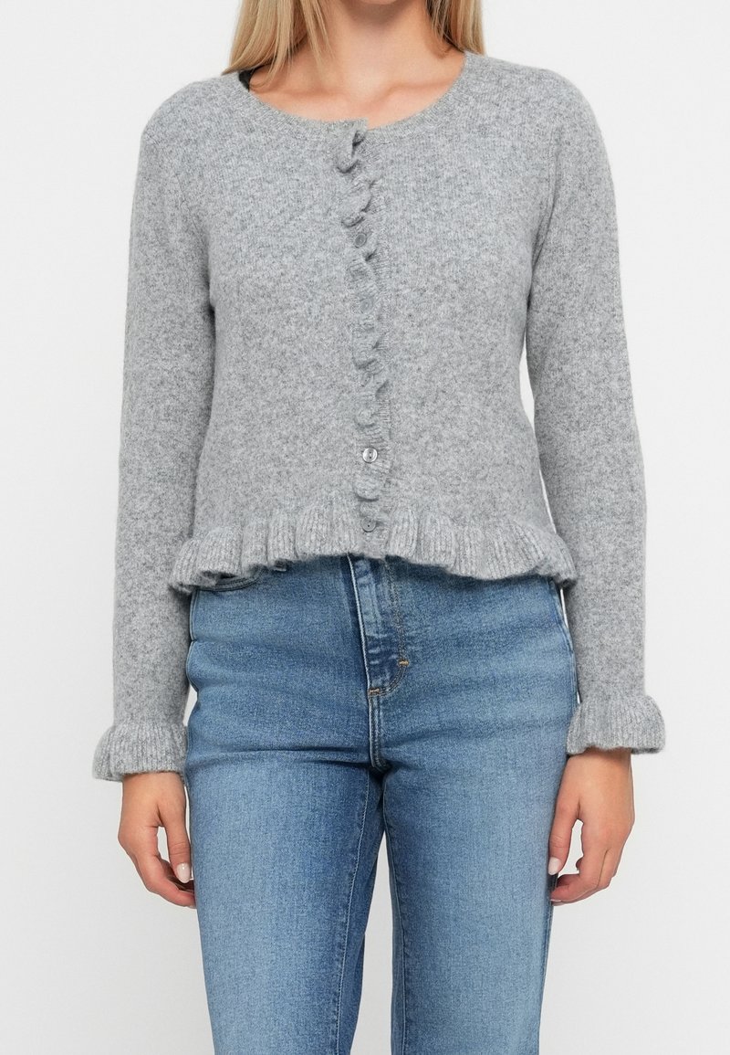 Femme portant un cardigan gris clair boutonné avec des bords volantés, assorti à un jean bleu taille haute sur un fond uni.
