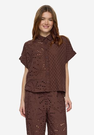 Nainen, jolla on yllään ruskea napitettava paita ja sitä vastaavat housut, joissa on kukkakuviolliset leikkaukset, seisoo valkoista taustaa vasten ja hymyilee hieman.