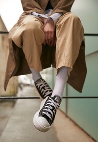 Baskets en toile noire avec semelles en caoutchouc blanches, bouts ronds et lacets blancs. Portées avec un pantalon crème et des chaussettes blanches.