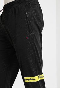 Svarta träningbyxor med en texturerad yta, med ett upprepande logomönster och en gul rand med text vid nederkanten.