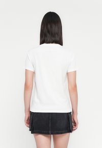 Woman with straight dark hair wearing a plain white short-sleeve t-shirt and a black denim mini skirt, facing away on a white background.
