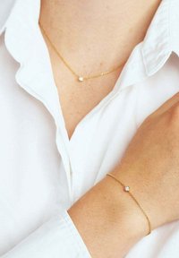 Delikat gyllene kedja med hänge och matchande armband, var och en med en liten rund klar ädelsten, bärs med en vit skjorta.