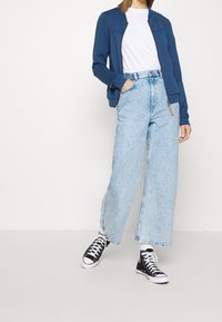 Blå bomberjacka, vit T-shirt och ljusblå vida jeans. Svarta sneakers med vita sulor. Avslappnad outfit med texturerad tyg.