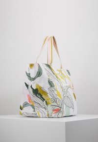 Sac fourre-tout en toile avec un motif floral coloré en verts, jaunes et roses, doté de poignées doubles rayées. Forme rectangulaire, texture lisse.