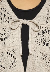 Cardigan de punto con frente abierto, en un color beige claro, con patrones de encaje y un cierre de lazo en el escote, usado sobre una blusa negra.