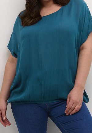 Mujer con una blusa suelta de manga corta color azul verdoso y vaqueros azules, de pie con una mano cerca de la cadera y la otra relajada.
