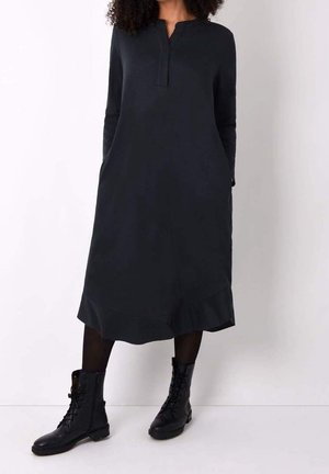 Kvinde iført en sort midi-kjole med lange ærmer og sidelommer, kombineret med sorte snørestøvler i militærstil på en ensfarvet hvid baggrund.