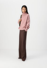 Rosa flauschiger Rollkragenpullover, weite braune Hose und eine grüne Umhängetasche. Das Outfit zeichnet sich durch einen lässigen Schnitt und weiche Texturen aus.