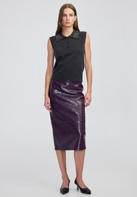 Kobieta w bezrękawnikowym ciemnoszarym swetrze z ozdobnym kołnierzem, błyszczącej fioletowej spódnicy do kolan o teksturowanej powierzchni oraz czarnych butach na obcasie.