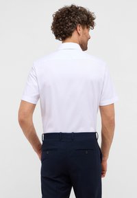 Camicia bianca a maniche corte con colletto classico, texture liscia e un retro con yoke, abbinata a pantaloni navali con due tasche posteriori.
