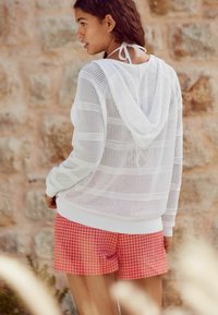 Witte, opengeweven hoodie met een trekkoord, gecombineerd met rood-witte geblokte shorts. Schone lijnen en een lichte textuur.