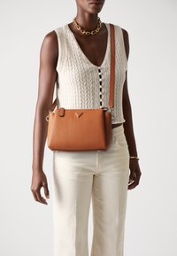 Sac en cuir texturé marron en bandoulière avec un logo triangulaire, complété par une longue sangle et des détails en matériel doré.