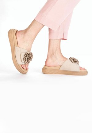 Sandales à glisser beiges avec une semelle tan texturée, ornées d'un accent floral sur la bride. Matériau lisse et design à bout ouvert.