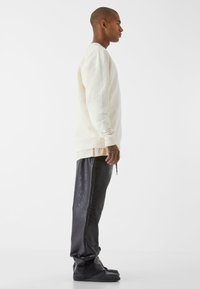 Felpa crema con polsini e orlo a coste, indossata sopra una camicia beige chiara. Pantaloni neri in tessuto strutturato con una vestibilità ampia e dettagli con coulisse.