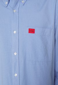 Camisa azul claro de botonadura hecha de algodón, con un bolsillo en el pecho que lleva una etiqueta roja "HUGO" y botones de plástico transparente. Textura suave, diseño casual.